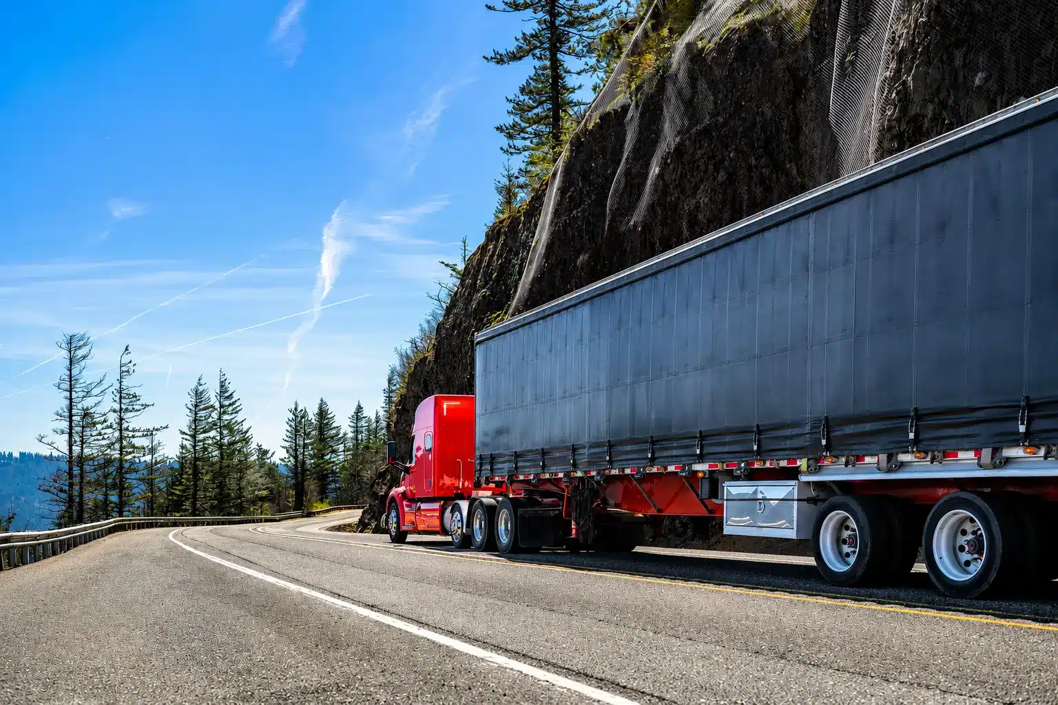 A red semi-truck with a black trailer, possibly hauling a flatbed truck load coordinated by RCT Logistics, drives on a winding mountain road lined with trees and a rocky cliff under a clear blue sky.