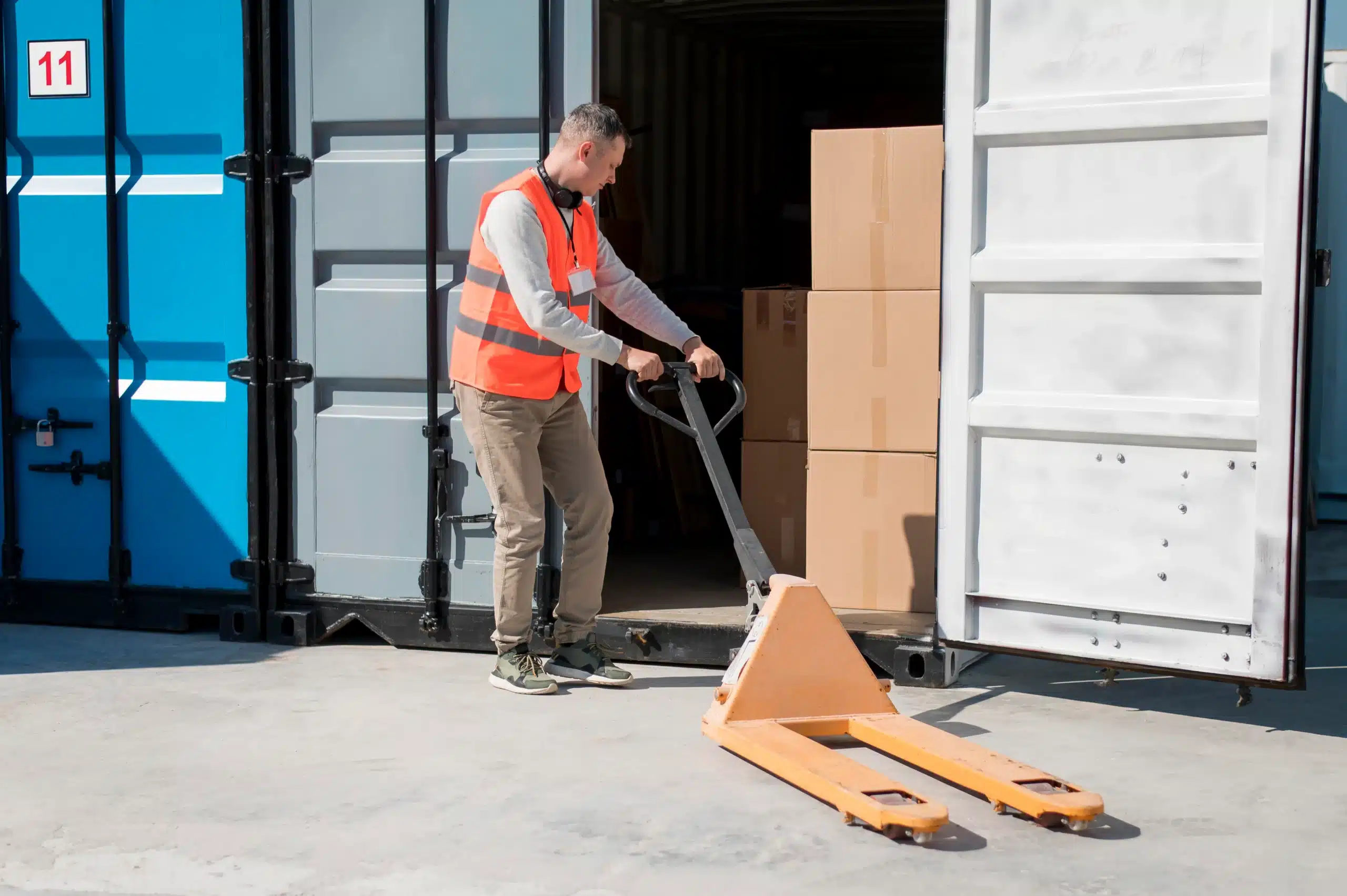 A worker in a safety vest moves a pallet jack near open shipping containers with stacked cardboard boxes inside, preparing for a flatbed truck load.