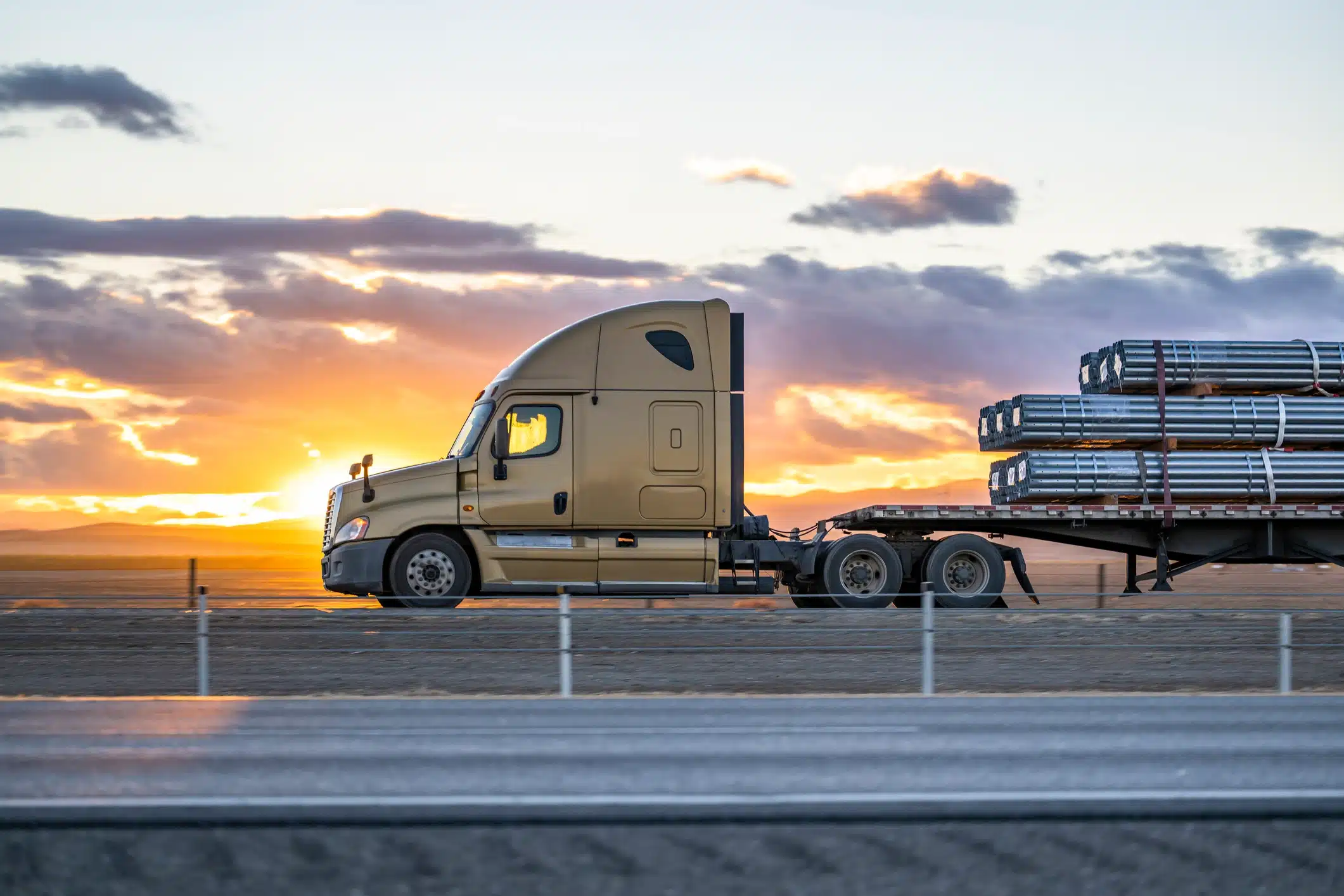 A semi-truck hauling long metal beams on a flatbed trailer, arranged by a West Michigan freight broker, drives along a road at sunset, with mountains in the background.