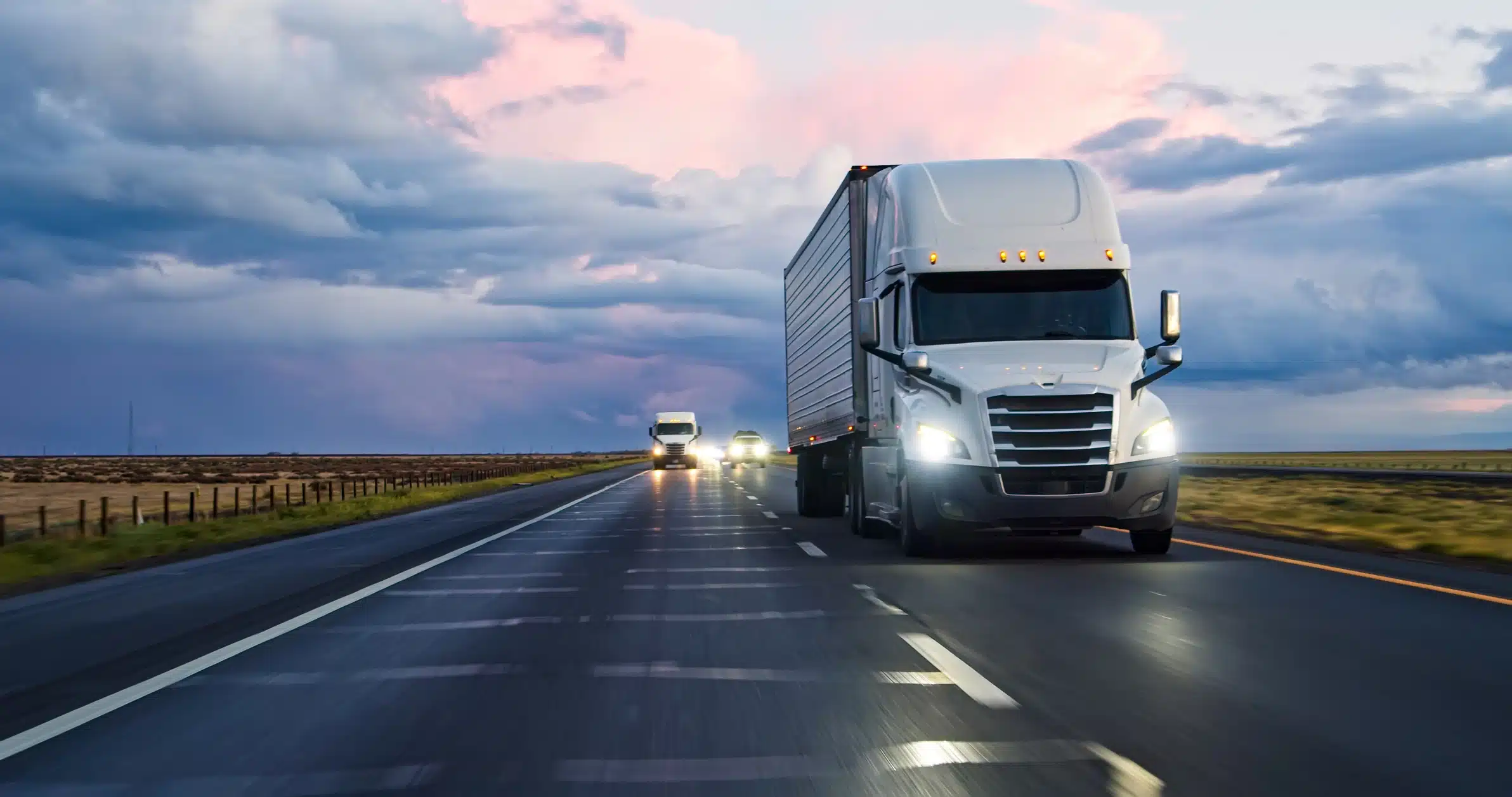 A white semi-truck with a dry van trailer drives down a highway at dusk, followed by other trucks, with cloudy skies and open fields on either side of the road.